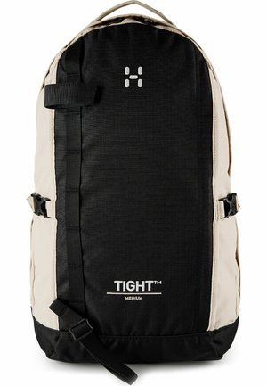 Zaino di dimensioni medie nero e beige con cinturino frontale, fibbie laterali, maniglia superiore e marchio "TIGHT™" sul pannello frontale.