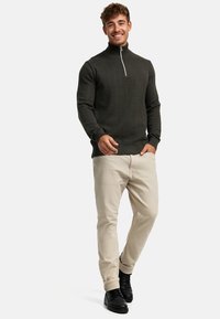 Mørkegrønn zip-neck genser med et teksturert herringbone-mønster, kombinert med lyse beige bukser og svarte snøresko mot en ensfarget bakgrunn.