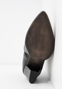Botte noire en cuir pointue avec une semelle texturée et un talon bloc, inclinée contre un fond blanc en angle.