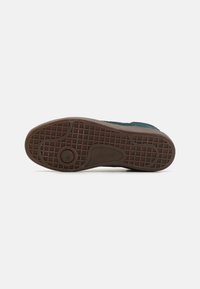 Zapato con una parte superior de ante verde oscuro texturizado y suela de goma marrón con un patrón de cuadrícula y un acento de logo circular.