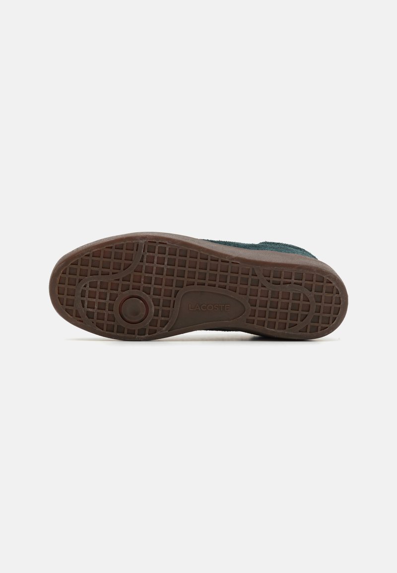 Zapato con una parte superior de ante verde oscuro texturizado y suela de goma marrón con un patrón de cuadrícula y un acento de logo circular.