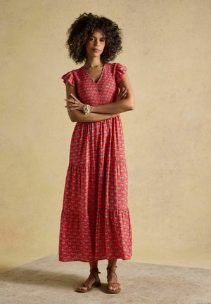 Woman with curly hair wearing a red floral maxi dress with flutter sleeves and tan ankle-tie sandals, standing with arms crossed.