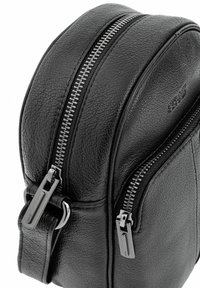 Czarna skórzana torba na ramię o okrągłym kształcie, z dwoma kontrastującymi srebrnymi zamkami błyskawicznymi, teksturowaną powierzchnią i regulowanym paskiem.