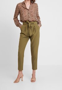 Brun blommig skjorta med knappar och långa ärmar, tillsammans med högmidjade olivgröna byxor med knytbälte och sidofickor. Ljusfärgade klackmules.