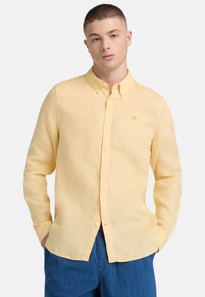 Junger Mann mit einem blassgelben Hemd und blauer Jeans, stehend mit den Händen in den Taschen vor einem einfarbigen hellen Hintergrund.