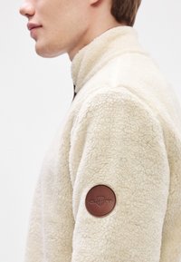 Cremefarvet fleecejakke med høj krave, struktureret stof og et brunt lædermærke med logo på venstre ærme.