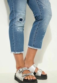 Jeans de mezclilla azules cortos con dobladillos deshilachados, con detalles de rodillas desgastadas y salpicaduras de pintura blanca, combinados con sandalias plateadas.