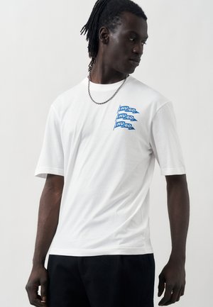 Άνδρας με κοτσιδάκια φοράει λευκό μπλουζάκι με τρεις μπλε σημαίες που γράφουν "HUGO" και ασημένιο αλυσίδα, κοιτάζει προς τα αριστερά του.