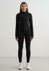 Černá sportovní bunda se zapínáním na zip, přiléhavý střih a dlouhé rukávy, doplněná o černé legíny a bílé tenisky.