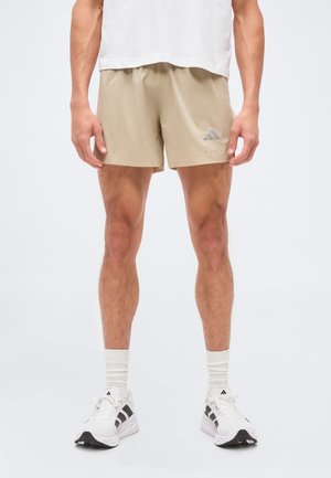 Άνδρας φορώντας μπεζ σορτς Adidas, λευκές κάλτσες και λευκά αθλητικά παπούτσια Adidas, στέκεται μπροστά σε ένα μονόχρωμο λευκό φόντο.