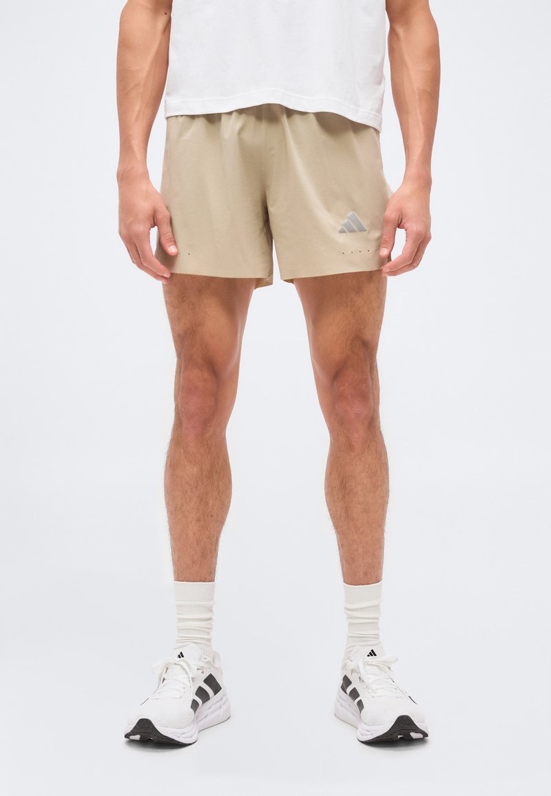 Άνδρας φορώντας μπεζ σορτς Adidas, λευκές κάλτσες και λευκά αθλητικά παπούτσια Adidas, στέκεται μπροστά σε ένα μονόχρωμο λευκό φόντο.