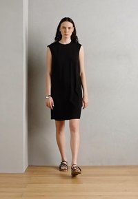 Robe noire sans manches en jersey, coupe droite, longueur genou, avec poches latérales. Portée avec des sandales noires à lanières sur un sol en bois.
