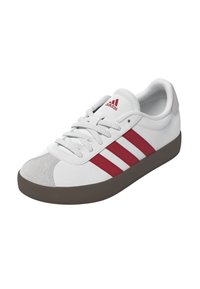 Zapatilla deportiva blanca con rayas rojas, parte superior sintética texturizada, puntera gris y suela de goma marrón. Presenta cordones tradicionales y un logo.