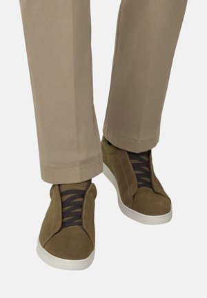Beige broek gecombineerd met olijfgroene suède sneakers met zwarte elastische veters en witte rubberen zolen op een witte achtergrond.
