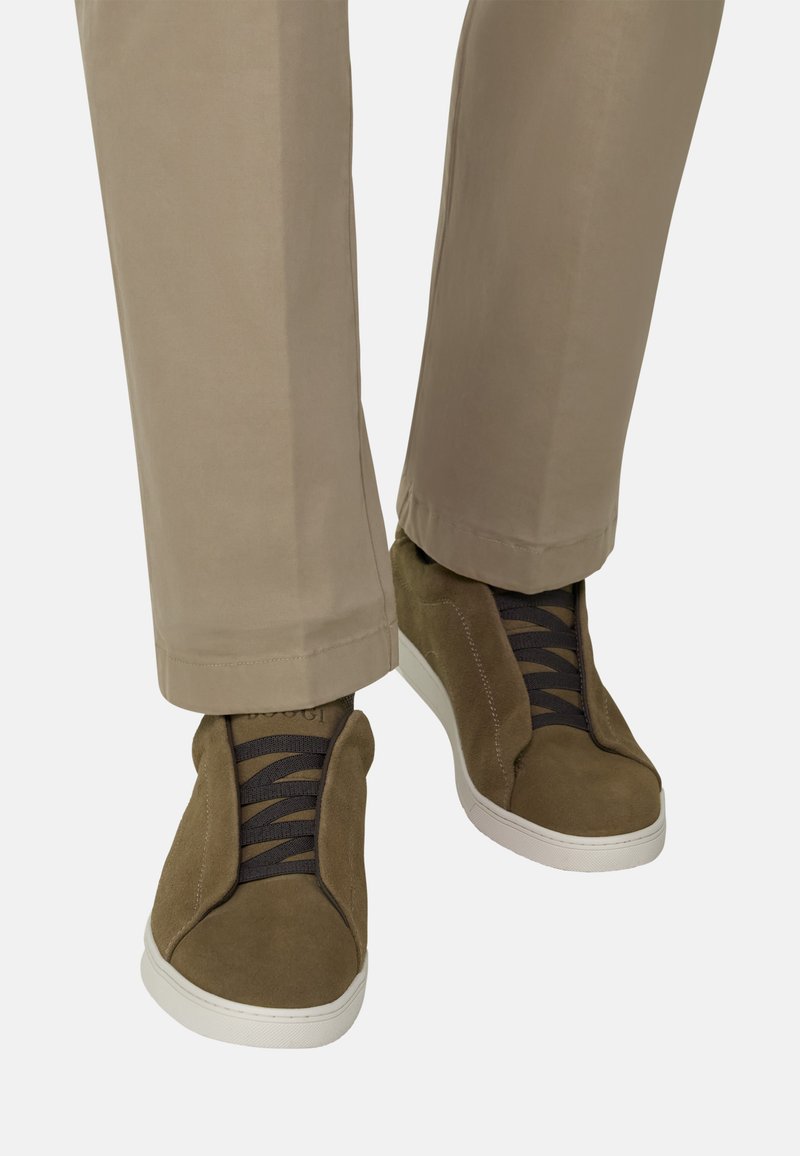 Pantalon beige associé à des baskets en daim vert olive avec des lacets élastiques noirs et des semelles en caoutchouc blanches sur fond blanc.