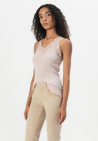 Top de alças em rosa claro com detalhes em renda nos ombros e no decote, combinado com calças ajustadas bege com bainha em formato de onda e costura decorativa.