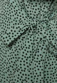 Robe en tissu vert avec de petits motifs floraux noirs, taille cintrée avec un détail en nœud, texture lisse et léger éclat.