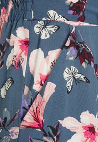 Tissu bleu avec imprimé floral rose et blanc ainsi que des papillons, montrant une ceinture froncée et un détail de poche latérale.