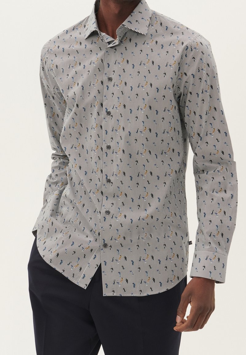 Homme portant une chemise blanche boutonnée avec de fines rayures noires et de petits motifs abstraits multicolores, assortie d’un pantalon foncé.