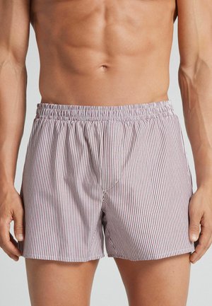 Torace maschile in forma che indossa boxer a vita elastica con sottili righe verticali rosse e bianche, su uno sfondo chiaro uniforme.