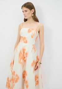 Kermadress med ett orange abstrakt mönster, som har tunna band och en åtsittande bodice som övergår i en utsvängd kjol. Slät tygtextur.