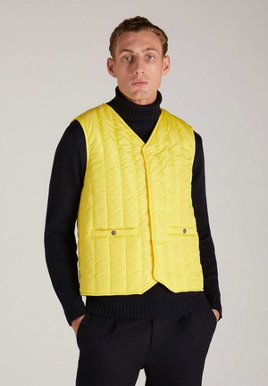 Gelbe gesteppte Weste mit V-Ausschnitt, zwei Fronttaschen und silbernen Hardware-Akzenten. Getragen über einem schwarzen Rollkragenpullover.