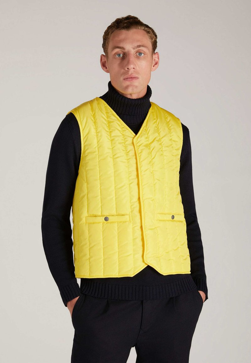 Gelbe gesteppte Weste mit V-Ausschnitt, zwei Fronttaschen und silbernen Hardware-Akzenten. Getragen über einem schwarzen Rollkragenpullover.