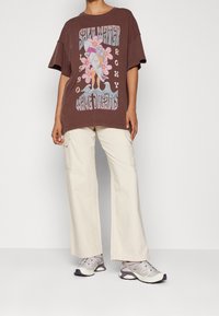 Camiseta gráfica marrón con diseños de tabla de surf y flores, combinada con pantalones cargo de color claro y zapatillas deportivas grises.