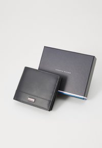 Portefeuille en cuir noir avec un détail de logo en argent, finition texturée et forme rectangulaire. Accompagné d'une boîte-cadeau bleu marine.
