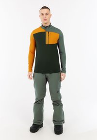 Groene en oranje longsleeve-shirt met een halve rits en borstzak, gecombineerd met groene broek en zwarte laarzen. Gladde stoftextuur.