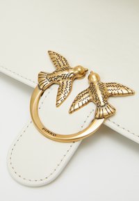 Accessorio in pelle bianca con una hardware circolare in oro che presenta due uccelli finemente progettati l'uno di fronte all'altro. Accenti testurizzati visibili.