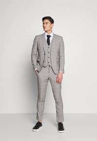 Grau kariertes dreiteiliges Anzugset mit einer taillierten Jacke, Weste und schmal geschnittenen Hosen, kombiniert mit einem weißen Hemd und einer schwarzen Krawatte sowie schwarzen Sneakern.