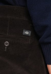 Pantalon en velours côtelé marron avec une étiquette portant un emblème, une fermeture à bouton et une surface texturée, associé à un pull en tricot bleu foncé.