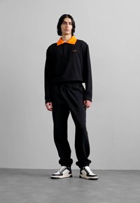 Sort langærmet polo med en orange krave, parret med sorte sweatpants og sort-hvide sneakers, med en afslappet pasform og snoretræk i taljen.