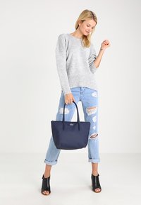 Navy leather tote bag with two handles, topped with a gold logo. Worn with a gray sweater and distressed light blue jeans.