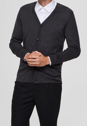 Personne portant un cardigan boutonné gris foncé par-dessus une chemise blanche à col, avec un pantalon noir, mains jointes devant.