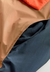 O geantă dintr-un material texturat de culoare maro și bleumarin, cu un capac portocaliu, parțial deschis pentru a dezvălui o căptușeală din fleece de culoare închisă în interior.