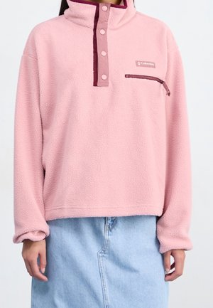 Personne portant un pull polaire Columbia rose clair avec des boutons-pression et une poche poitrine zippée, associé à une jupe en jean bleu clair.