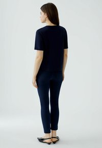 Marineblauwe T-shirt met korte mouwen, gecombineerd met skinny donkere jeans. Model gezien van achteren. Zwarte slingback-schoenen. Glad stoftextuur. Minimalistisch ontwerp.
