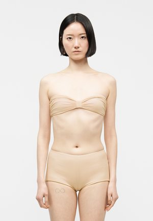 Femme aux cheveux courts et noirs portant un haut bandeau beige et un short taille haute assorti, debout devant un fond blanc uni.