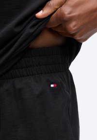 Χέρι σηκώνει μαύρο πουκάμισο για να αποκαλύψει τη μέση μαύρου ελαστικού παντελονιού με μικρό ορθογώνιο λογότυπο σε κόκκινο, λευκό και μπλε χρώμα.