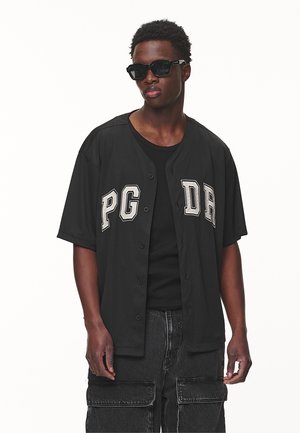 Black button-up jersey with "PG" and "DR" printed in cream, paired with a black tank top and loose black cargo pants. Black sunglasses worn.