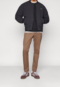 Svart bomberjacka med ribbade muddar och nederkant, lagt över en mörk sweatshirt. Bruna tapered chinos och bruna sneakers med vita detaljer.