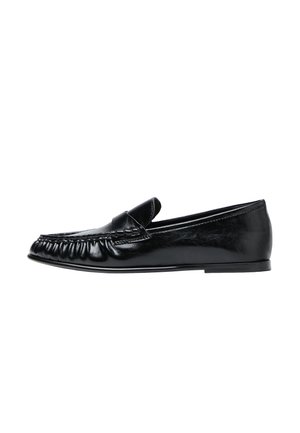 Mocassin noir en cuir à enfiler avec des détails cousus et une semelle plate, présenté de profil sur fond blanc.