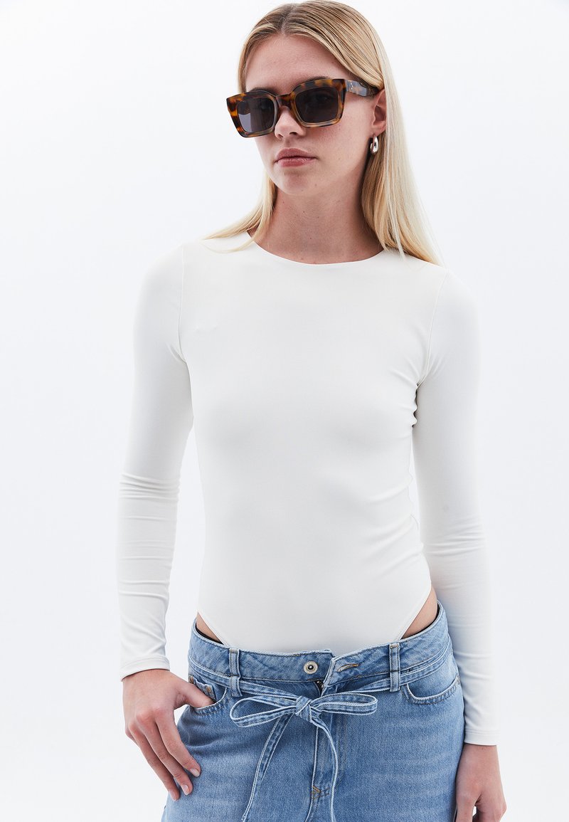 Weißer Langarm-Body aus glattem Stoff, kombiniert mit einem hellblauen Denim-Rock, der ein Bindeband-Detail an der Taille hat.