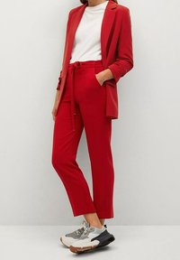 Žena v červeném blejzru a odpovídajících kalhotách, s bílým tričkem a šedo-bílými teniskami, stojící na jednobarevném světlém pozadí.