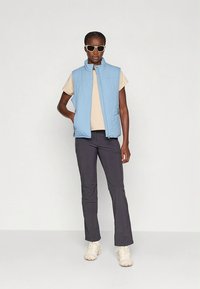 Modro svetla brezrokavnik čez bež majico, kombiniran s temnimi hlačami in kremnimi supergami. Model nosi sončna očala; gladka tekstura tkanine.