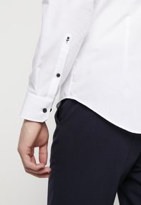 Camicia bianca a maniche lunghe, con bottoni neri e dettaglio di rosa nero ricamato sul polsino, abbinata a pantaloni blu scuro.