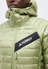 Κοντινό πλάνο ενός ατόμου που φοράει ένα ανοιχτό πράσινο μπουφάν καπιτονέ με μαύρο διαγώνιο μοτίβο και λευκό λογότυπο "TERREX" στο στήθος.