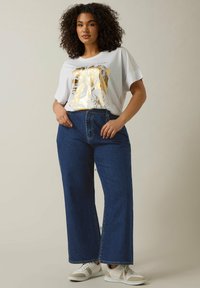 Camiseta blanca con estampado floral dorado, combinada con jeans de mezclilla azul de tiro alto y holgados, y zapatillas blancas con acentos beige.